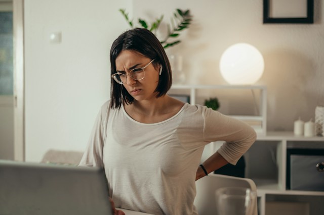 Woman at her desk at home holding her lower back, tired after a long day Woman at her desk at home holding her lower back, tired after a long day